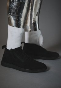 Botas de tobillo de ante negro con detalle de cremallera, suela texturizada negra y combinadas con calcetines blancos con canalé. Piernas metálicas brillantes al fondo.