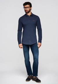 Homme portant une chemise bleu marine à manches longues boutonnée, un jean bleu foncé, des lunettes noires et des chaussures marron foncé à lacets, debout devant un fond uni.