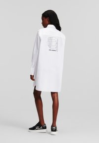 Abito camicia bianco con maniche lunghe, caratterizzato da un design grafico sulla schiena. Abbinato a sneakers nere con accenti scintillanti.