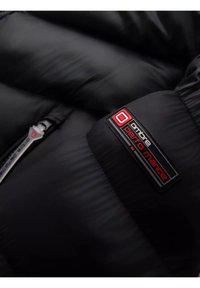Giacca imbottita nera in tessuto lucido. Presenta un logo prominente in rosso e nero e una zip argento.