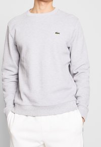 Uomo che indossa un maglione grigio chiaro a girocollo con logo di coccodrillo verde sul petto e pantaloni bianchi, mani in tasca.