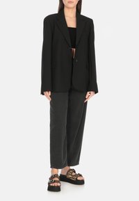 Blazer nero oversize con due bottoni frontali, abbinato a pantaloni neri a vestibilità rilassata e sandali neri e beige con fantasia.