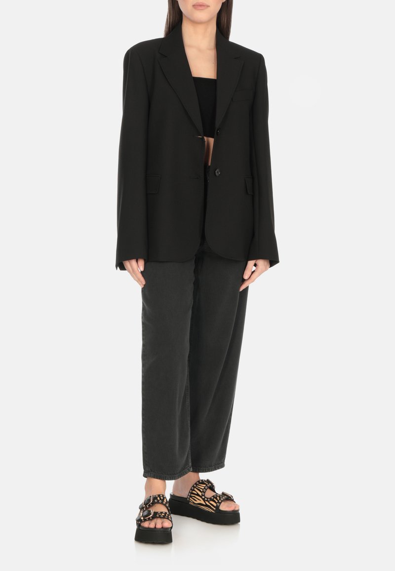 Blazer nero oversize con due bottoni frontali, abbinato a pantaloni neri a vestibilità rilassata e sandali neri e beige con fantasia.