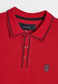 Czerwony golf polo z teksturowanym splotem, zapięcie na trzy guziki, dwie czarne paski na kołnierzyku oraz wyszywany logo lwa po lewej stronie klatki piersiowej.