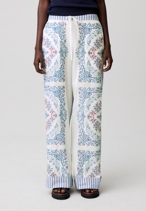 Pantaloni bianchi a gamba larga con motivi intricati blu e rossi, polsini a righe e vita con coulisse, indossati con sandali scuri e top blu navy.