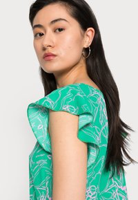 Blusa verde com mangas em babados, apresentando um padrão floral em rosa. Textura suave, corte solto e detalhe visível nos ombros.