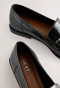 Mocassins noirs en cuir à enfiler avec petits talons et semelles intérieures rembourrées marron, photographiés sur un fond clair.