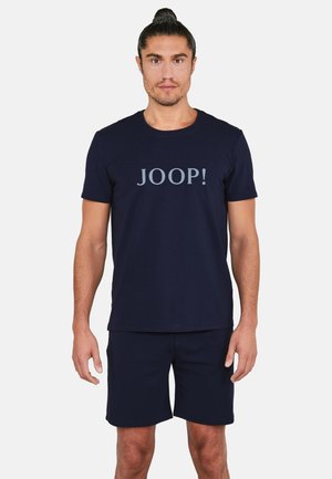 Mann mit zusammengebundenen Haaren, trägt ein dunkelblaues T-Shirt mit dem Schriftzug "JOOP!" und passende dunkelblaue Shorts, steht vor einem schlichten hellen Hintergrund.
