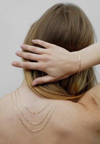 Gold layered chain necklace drapes across bare skin; features three strands with a textured finish. Fine gold bracelet on wrist.