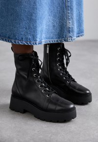 Bottes en cuir noir à lacets avec une semelle en caoutchouc épaisse, dotées de bouts ronds, d'œillets en métal et d'une fermeture éclair sur le côté.