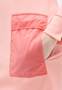 Χέρι στην πλαϊνή τσέπη ενός ρουχισμού ανοιχτού ροζ χρώματος με αντίθετη λεπτομέρεια τσέπης και μανσετών σε κοραλί απόχρωση.