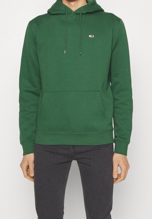 Mann trägt einen grünen Kapuzenpullover mit Kordelzügen und einer aufgesetzten Kängurutasche vorne, kombiniert mit dunkelgrauen Hosen.