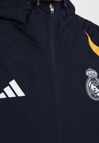 Marineblauw jack met ritssluiting en capuchon, met wit Adidas-logo aan de linkerkant en Real Madrid-embleem aan de rechterkant, met gele accenten op de schouder.