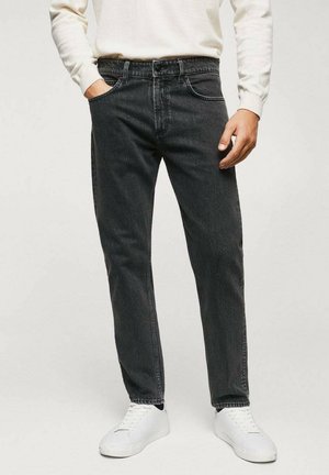 Mann trägt schwarze Jeans, weiße Sneaker und ein weißes langärmliges Hemd, mit einer Hand in der Tasche, vor einfarbigem Hintergrund.