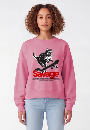 Jeune femme aux cheveux bouclés portant un sweat-shirt rose avec un motif représentant un chat en skateboard au-dessus du mot "Savage" en lettres rouges.