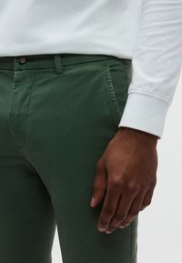 Στενά, σκούρα πράσινα βαμβακερά παντελόνια με μπροστινές τσέπες. Ορατό κουμπί στη μέση. Λευκό πουκάμισο με μακριά μανίκια εμφανίζεται εν μέρει στην κορυφή.