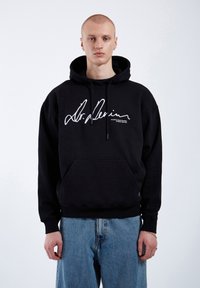 Black cotton hoodie with a drawstring hood, front pocket, and large white printed signature on the chest, paired with light blue denim jeans.