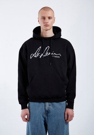 Black cotton hoodie with a drawstring hood, front pocket, and large white printed signature on the chest, paired with light blue denim jeans.