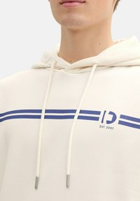 Sudadera crema con capucha, con rayas azules y texto de logo. Hecha de material suave, destacada con cordones con puntas metálicas.