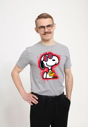Ung mand med briller og moustache iført en grå t-shirt med Snoopy med røde solbriller og Woodstock, stående med den ene hånd på hoften.