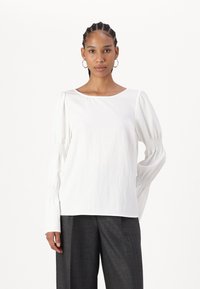 Blusa bianca a maniche lunghe con una texture liscia, caratterizzata da dettagli smock sulle maniche e un colletto rotondo. Indossata con pantaloni grigi.