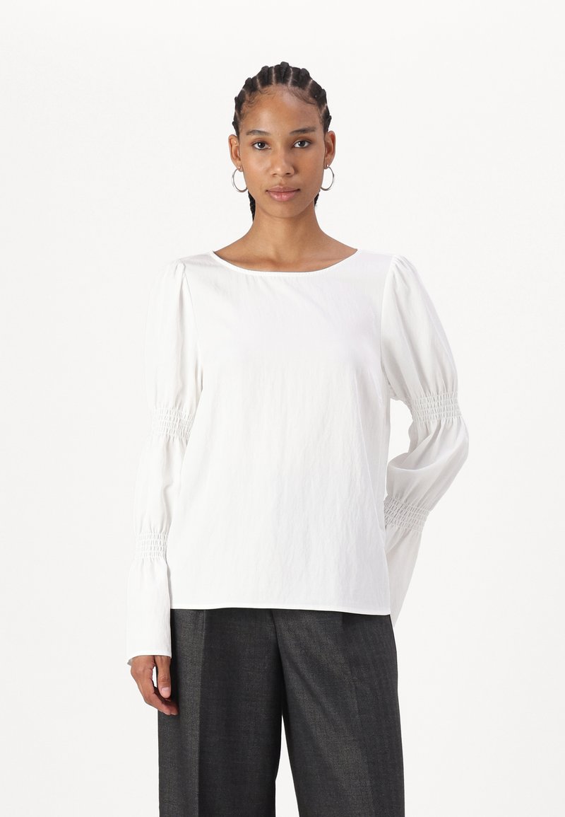 Blusa bianca a maniche lunghe con una texture liscia, caratterizzata da dettagli smock sulle maniche e un colletto rotondo. Indossata con pantaloni grigi.