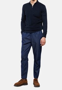 Maglione zip in maglia blu navy, pantaloni con coulisse blu navy e scarpe in suede marrone con suole testurizzate. L'insieme presenta colori e materiali coordinati.