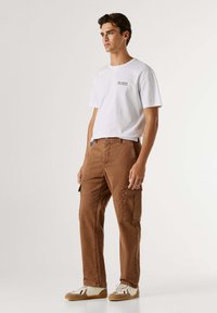 Bruine cargobroek met meerdere zakken, gecombineerd met een wit T-shirt met korte mouwen en een klein logo. Casual schoenen maken de outfit compleet.