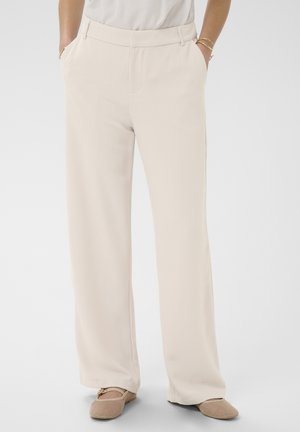 Frau trägt hochgeschnittene, weit geschnittene cremefarbene Hosen mit Seitentaschen und beige gewebte flache Schuhe, steht vor einem einfarbigen Hintergrund.
