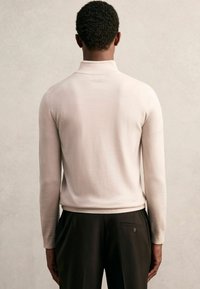 Helle beige Strickpullover mit hohem Kragen, langen Ärmeln und slim fit, kombiniert mit dunklen Hosen, die ein Rückentaschen-Detail aufweisen.