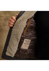 Interiér koženej bundy s béžovým umelým kožušinovým podšívaním, prešívanou hnedou tkaninou a označením. Obsahuje zipsovú kapsu s oranžovým ťaháčikom.