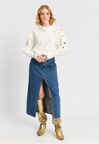 Naine kannab valget pikka varrukatoppi, millel on kuldsed nööbid varrukatel, eespool lõhiku denim seelikut ja kuldseid kuni põlveni ulatuvaid saapaid, seistes valge tausta ees.