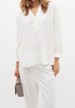 Kobieta ubrana w luźną, białą bluzkę z długim rękawem i spodnie, trzymająca złożoną torebkę kopertówkę koloru taupe przy boku.