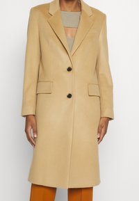 Cappotto beige in lana con revers a punta, due tasche frontali, chiusura con un singolo bottone nero, taglio dritto e texture morbida. Indossato sopra una maglietta chiara.