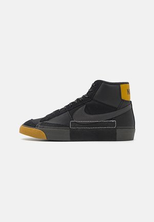 Czarny sneaker typu high-top z ciemnoszarym logo, żółtą łatką na pięcie, czarnymi sznurowadłami i brązowym teksturowanym czubkiem na białym tle.