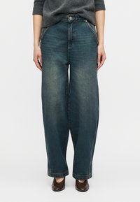 Άτομο που φοράει φαρδιά, ψηλόμεσα μπλε τζιν παντελόνια, σε συνδυασμό με γκρι πουλόβερ και καφέ παπούτσια με τακούνι, στέκεται μπροστά σε απλό φόντο.