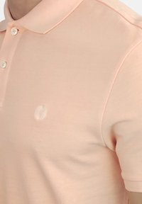 Polo de color melocotón claro hecho de algodón texturizado, con cuello clásico, tapeta de tres botones y un logo sutil en el lado izquierdo del pecho.