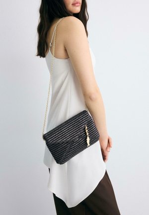 Ženska, oblečena v belo brezrokavnik in rjave hlače, nosi črno tkano torbico čez ramo z zlatim verižnim trakom in obeskom v obliki ribe.