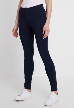 Mujer que lleva pantalones ajustados azul marino con bolsillos con cremallera y zapatillas blancas, de pie frente a un fondo claro y liso.