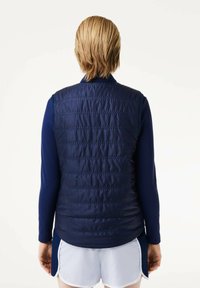 Chaleco acolchado azul marino con costuras horizontales y cuello alto, llevado sobre una camiseta de manga larga. Presenta una textura suave y brillante, adecuada para usar en capas.