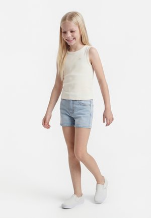 Simpatična djevojka s dugom plavom kosom koja nosi kremasti top bez rukava, svijetle traper kratke hlače i bijele slip-on cipele, hoda na bijeloj pozadini.