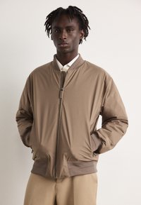 Bruine bomberjack van soepel materiaal, met een rits aan de voorkant, ribgebreide cuffs en een casual pasvorm, gecombineerd met een licht shirt eronder.