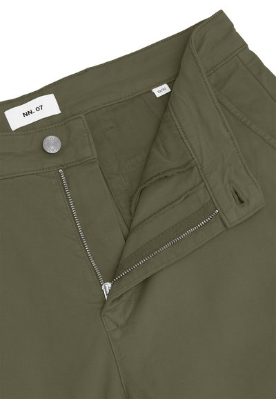 Pantalon vert olive avec fermeture éclair argentée partiellement ouverte, bouton en métal gravé "NN. 07" et étiquette de taille visible 32/32 à l'intérieur de la ceinture.