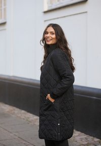 Black quilted long coat with a zippered pocket, ribbed collar, and side slits. Features minimalistic design, soft texture, and tailored fit.