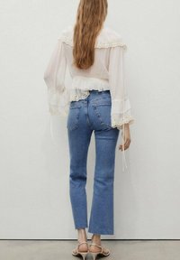 Bluză albă cu dantelă la margine, asociată cu blugi evazați de culoare albastră deschisă. Bluza are mâneci lungi, volanate și talie strânsă.