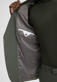 Blazer verde con una fodera interna grigia liscia, con patta della tasca bianca e dettaglio con bottone all'interno. Il tessuto esterno ha una finitura testurizzata.