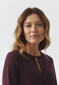 Blouse bordeaux avec un détail à encolure en porte-clé. Texture lisse et manches longues. Le modèle a des cheveux bruns ondulés et des yeux bleus.