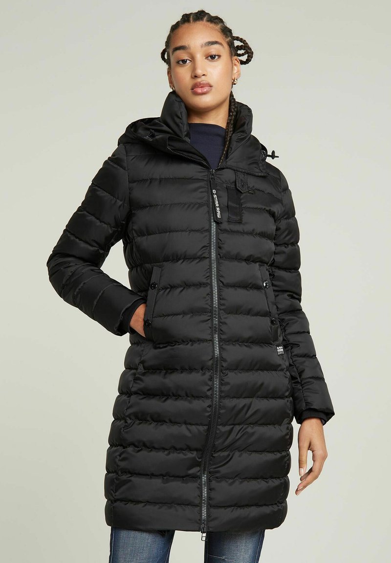 Schwarzer Puffer-Mantel mit einem Reißverschluss, Kapuze und gerippten Bündchen. Verfügt über vertikale Nähte und zwei Vordertaschen zur Aufbewahrung.