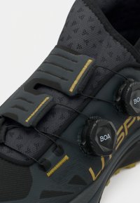 Czarny i szary but sportowy z siatkową cholewką, elastycznymi sznurówkami i dwoma regulowanymi pokrętłami BOA; posiada żółte przeszycia i teksturowaną podeszwę.