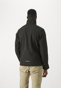 Czarna kurtka softshell z wysokim kołnierzem i odblaskowym akcentem z tyłu, w połączeniu z khaki spodniami cargo. Gładka faktura.